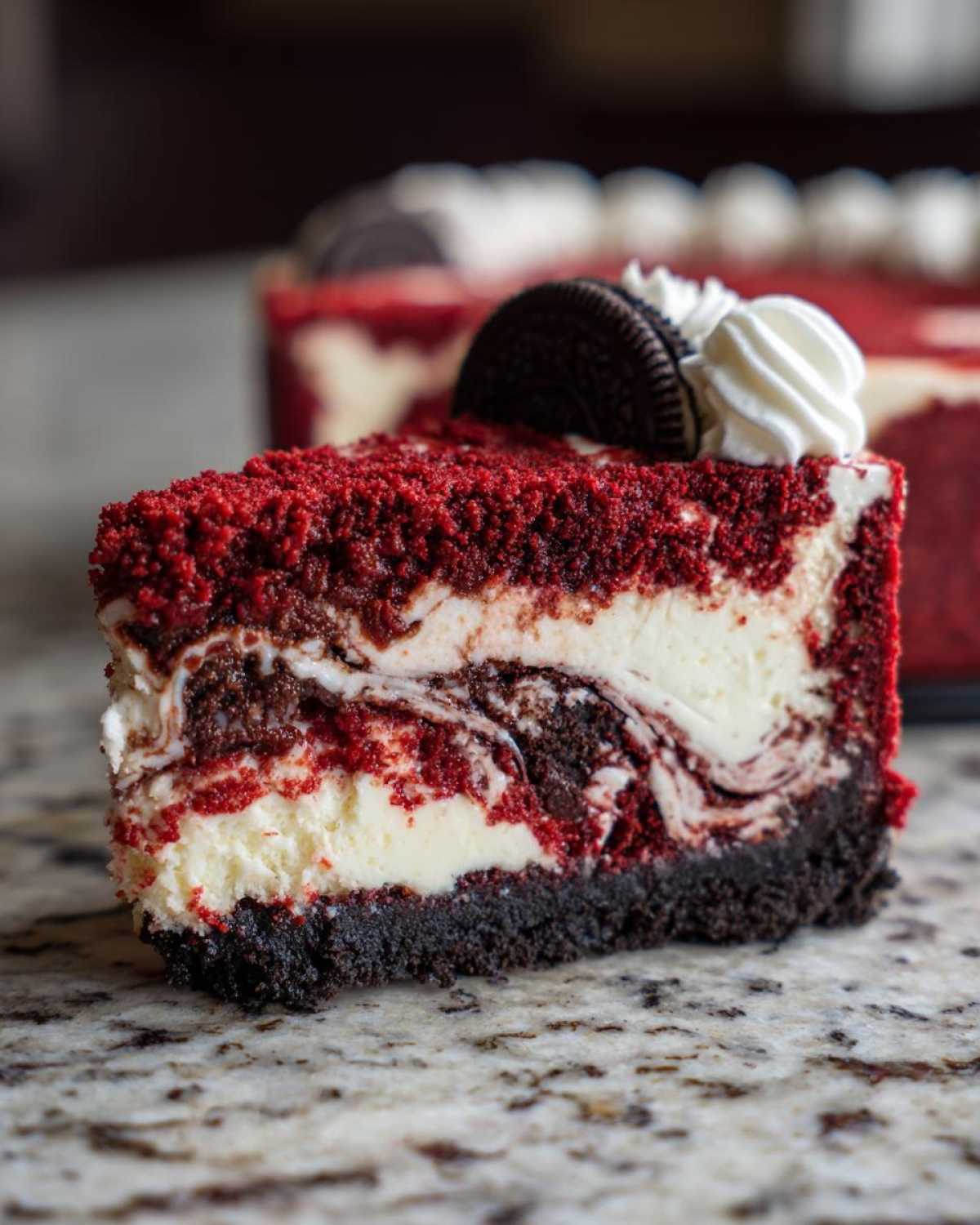 Close-up of a slice of red velvet cheesecake showing marbled layers, topped with whipped cream and a chocolate sandwich cookie.