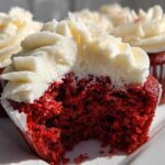 A close-up of a red velvet cupcakes cut in half, showing the moist red cake and thick swirl of white cream cheese frosting.