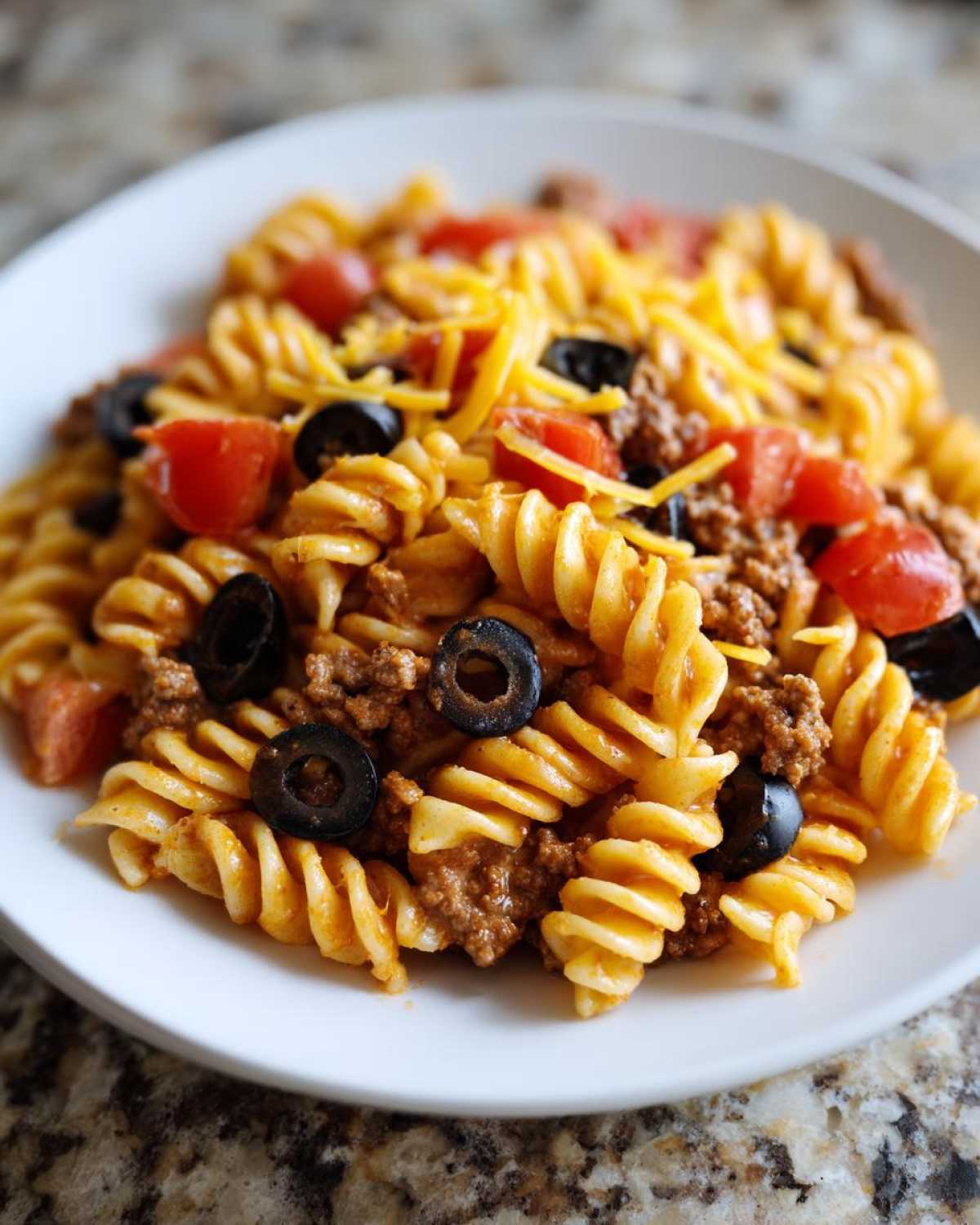 Close-up of a serving of taco pasta salad featuring rotini pasta, seasoned ground beef, black olives, tomatoes, and shredded cheese.