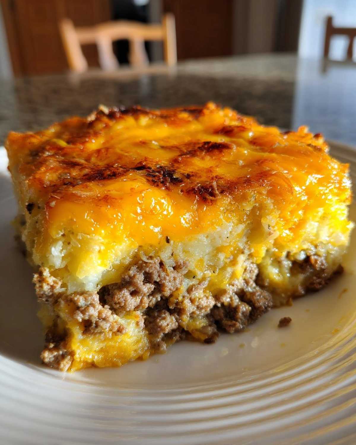 A close-up of a square serving of hamburger potato casserole featuring a layer of ground beef topped with potatoes and a thick layer of melted, browned cheddar cheese.