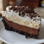 A decadent slice of layered mud pie featuring a dark crust, chocolate mousse, vanilla layer, and whipped cream topped with chocolate shavings.