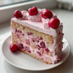 A close-up slice of moist cherry cake layered with pink cream and fresh cherries, topped with piped frosting and whole cherries.