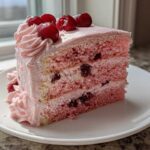 A perfect slice of pink layered Cherry cake with visible cherries inside and topped with fresh cherries and pink frosting.