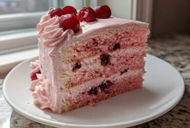 A perfect slice of pink layered Cherry cake with visible cherries inside and topped with fresh cherries and pink frosting.