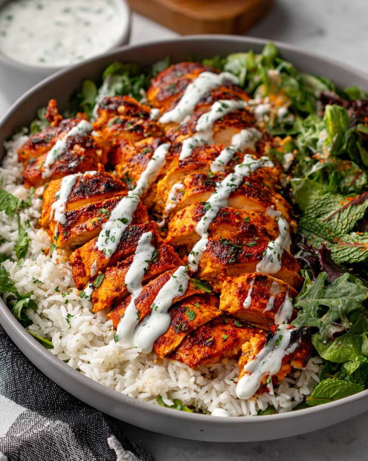 Close-up of sliced, spiced harissa chicken drizzled with a white yogurt sauce, served over white rice and mixed greens.