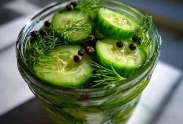 Close-up of sliced cucumbers, fresh dill, and black peppercorns ready for refrigerator pickles in a glass jar.