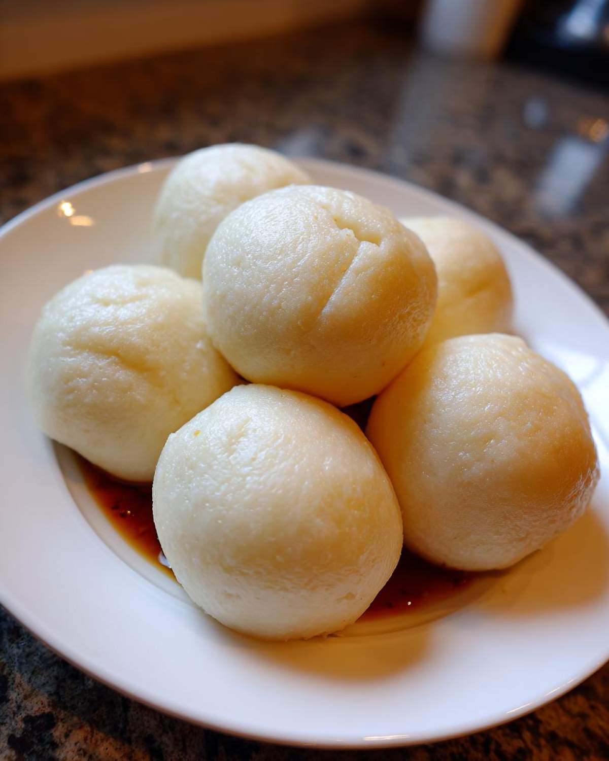Close-up of several smooth, pale yellow balls of fufu resting in a small pool of sauce on a white plate.