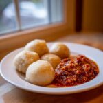 Five smooth, pale balls of fufu served on a white plate next to a rich, textured red pepper stew.
