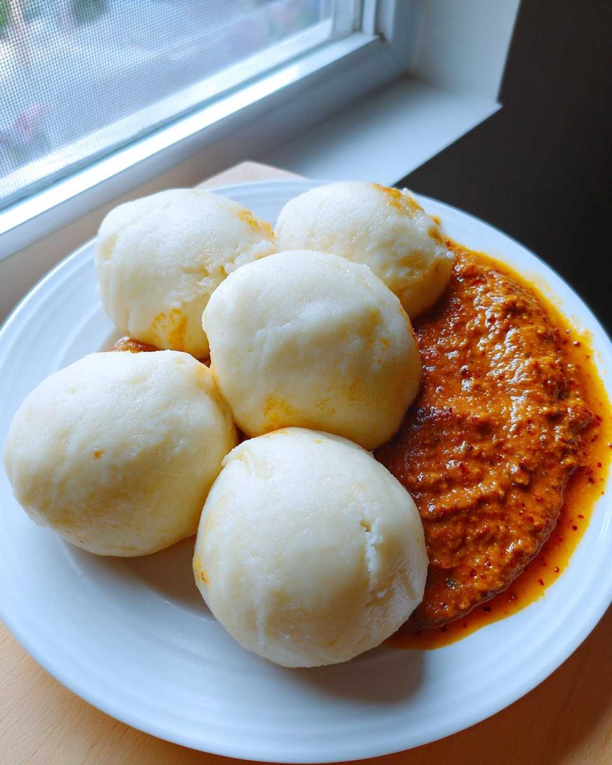 Five perfectly smooth balls of white fufu served on a white plate next to a rich, reddish-orange stew.