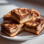 Close-up of stacked snickerdoodle blondies squares with a rich cinnamon swirl topping on a white plate.