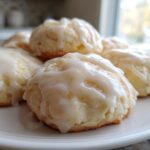 A close-up of several soft ricotta cookies piled on a white plate, generously topped with a thick white glaze.