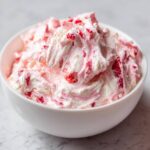 Close-up of creamy, swirled strawberry cheesecake dip mixed with visible strawberry pieces in a white bowl.