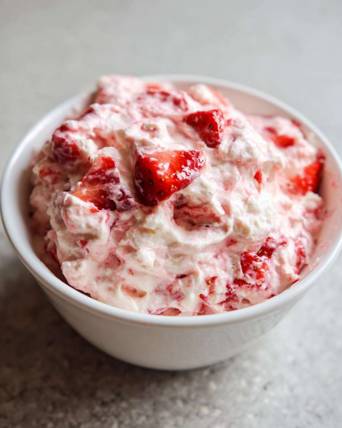 A close-up view of creamy, pink strawberry cheesecake dip mixed with chunks of fresh strawberries in a white bowl.