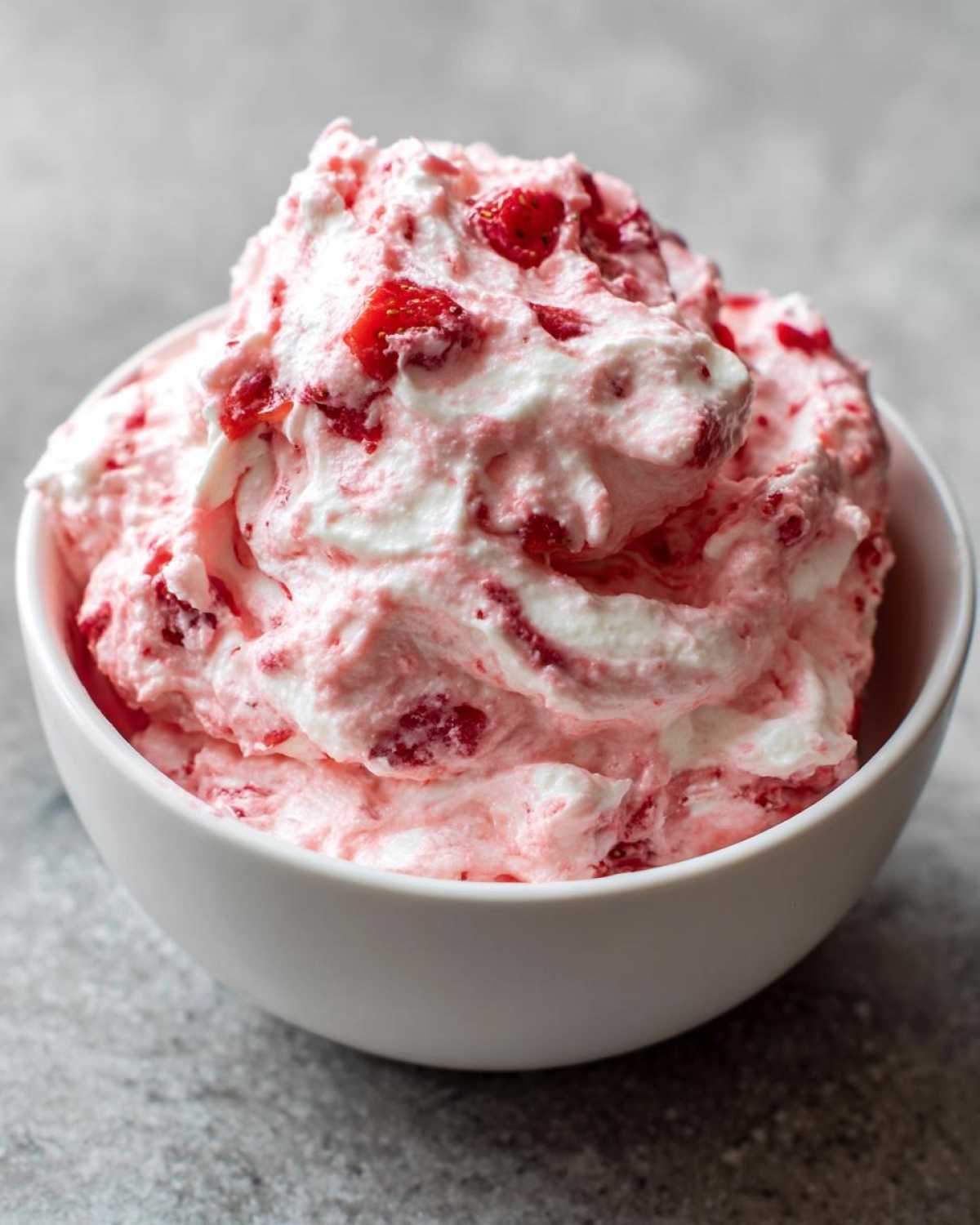 Close-up of fluffy, pink and white strawberry cheesecake dip mixed with strawberry pieces, served in a white bowl.