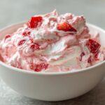 Close-up of light pink and white strawberry cheesecake dip topped with fresh strawberry chunks in a white bowl.