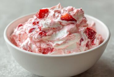 Close-up of light pink and white strawberry cheesecake dip topped with fresh strawberry chunks in a white bowl.