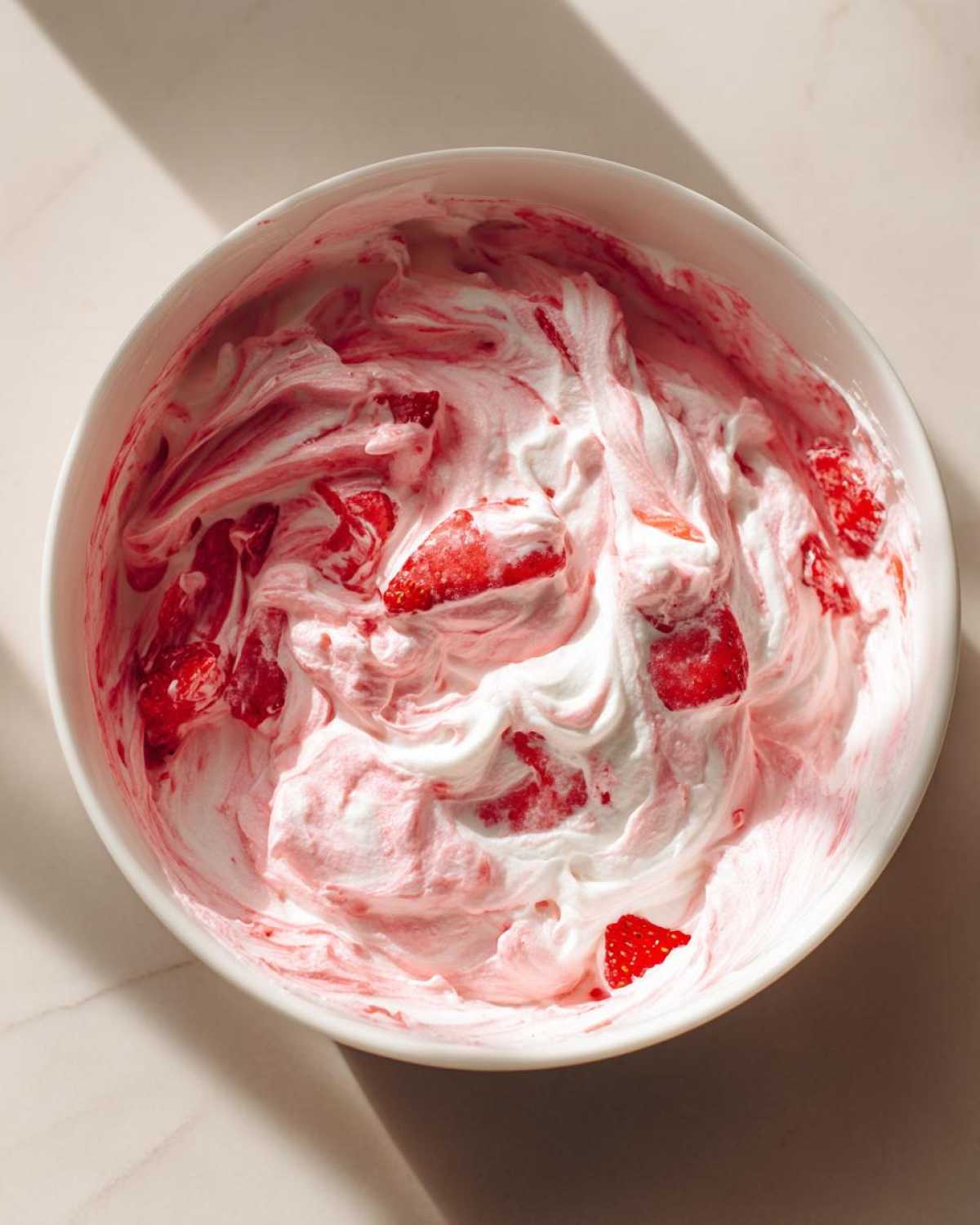 Overhead view of creamy strawberry cheesecake dip, swirled pink and white with chunks of fresh strawberries, in a white bowl.