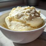 Close-up of creamy vanilla protein ice cream scooped high in a white bowl, brightly lit by sunlight.