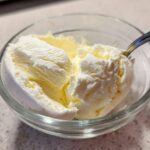 Close-up of creamy, pale yellow cottage cheese ice cream scoops in a clear glass bowl with a spoon.