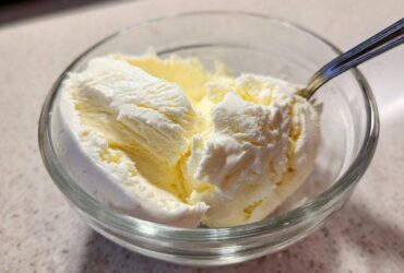 Close-up of creamy, pale yellow cottage cheese ice cream scoops in a clear glass bowl with a spoon.