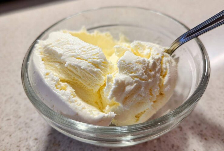 Close-up of creamy, pale yellow cottage cheese ice cream scoops in a clear glass bowl with a spoon.
