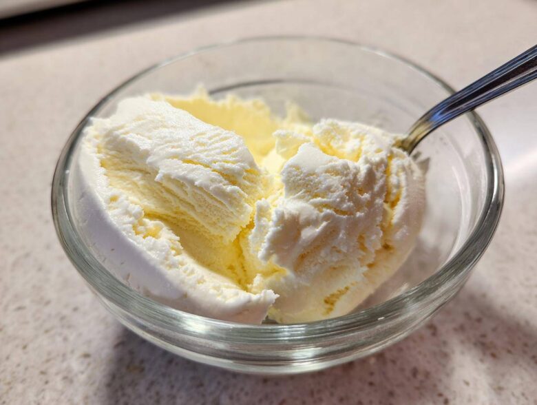 Close-up of creamy, pale yellow cottage cheese ice cream scoops in a clear glass bowl with a spoon.