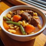 Close-up of a white bowl filled with rich vegetable beef soup, featuring chunks of beef, carrots, green beans, and elbow macaroni.