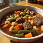 Close-up of a white bowl filled with rich vegetable beef soup featuring chunks of beef, carrots, green beans, and rotini pasta.