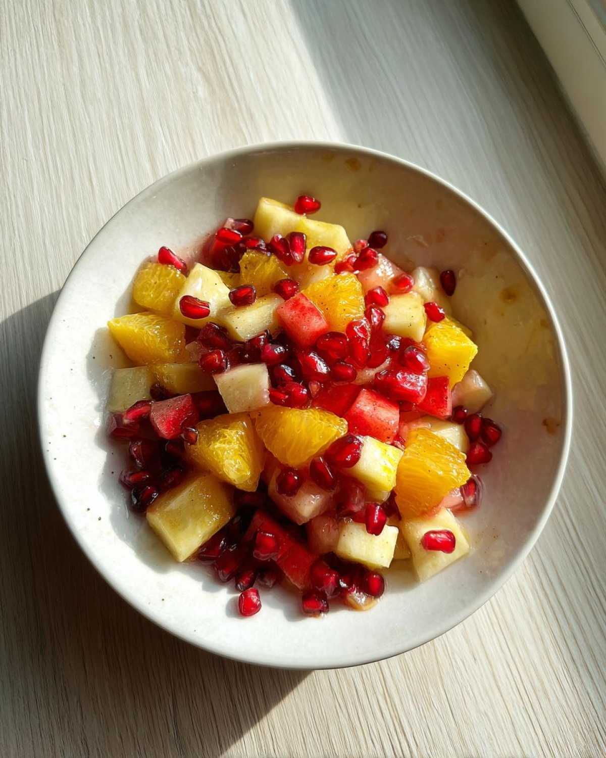A close-up of a bowl containing a vibrant winter fruit salad with orange segments, pineapple chunks, and bright red pomegranate seeds.