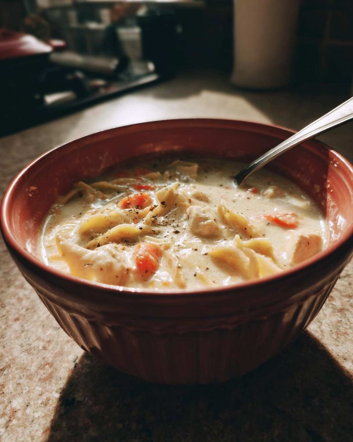 A close-up of creamy white chicken lasagna soup with noodles and carrots, seasoned with pepper, in a reddish bowl.