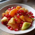 Close-up of a bright winter fruit salad featuring orange segments, apple chunks, and vibrant pomegranate seeds in a white bowl.