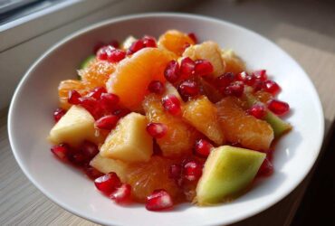 Close-up of a bright winter fruit salad featuring orange segments, apple chunks, and vibrant pomegranate seeds in a white bowl.