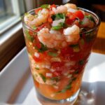 Close-up of a refreshing shrimp cocktail in a glass, featuring shrimp, diced tomatoes, onions, and cilantro in a tomato-based sauce.