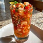 Close-up of a tall glass filled with zesty shrimp cocktail, featuring plump shrimp, tomatoes, cilantro, and a lime wedge garnish.