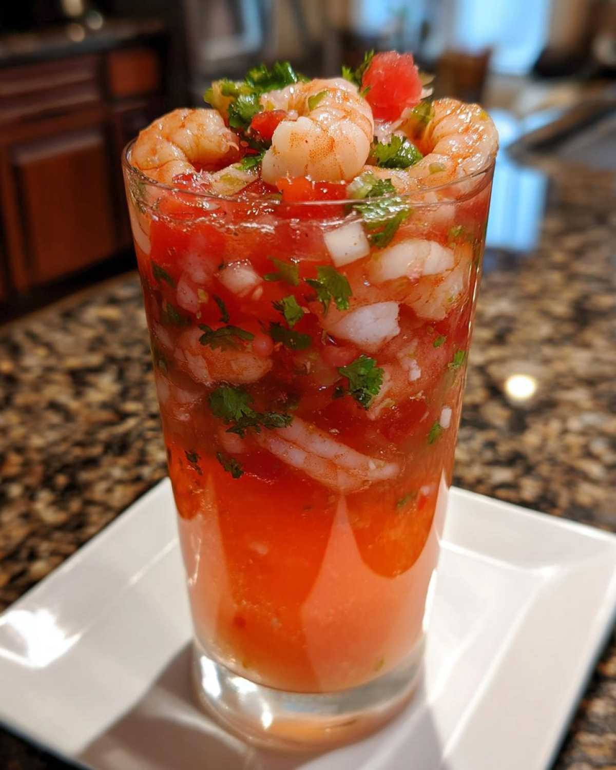 A tall glass filled with a vibrant, chilled shrimp cocktail mixed with diced tomatoes, onions, and cilantro.