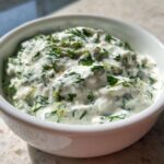 A white bowl filled with thick, creamy 5-Minute Greek Yogurt Ranch Dip, heavily speckled with fresh green herbs like dill and parsley.