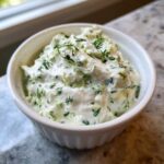 A close-up of creamy 5-Minute Greek Yogurt Ranch Dip, flecked with green herbs like dill, served in a small white ramekin.
