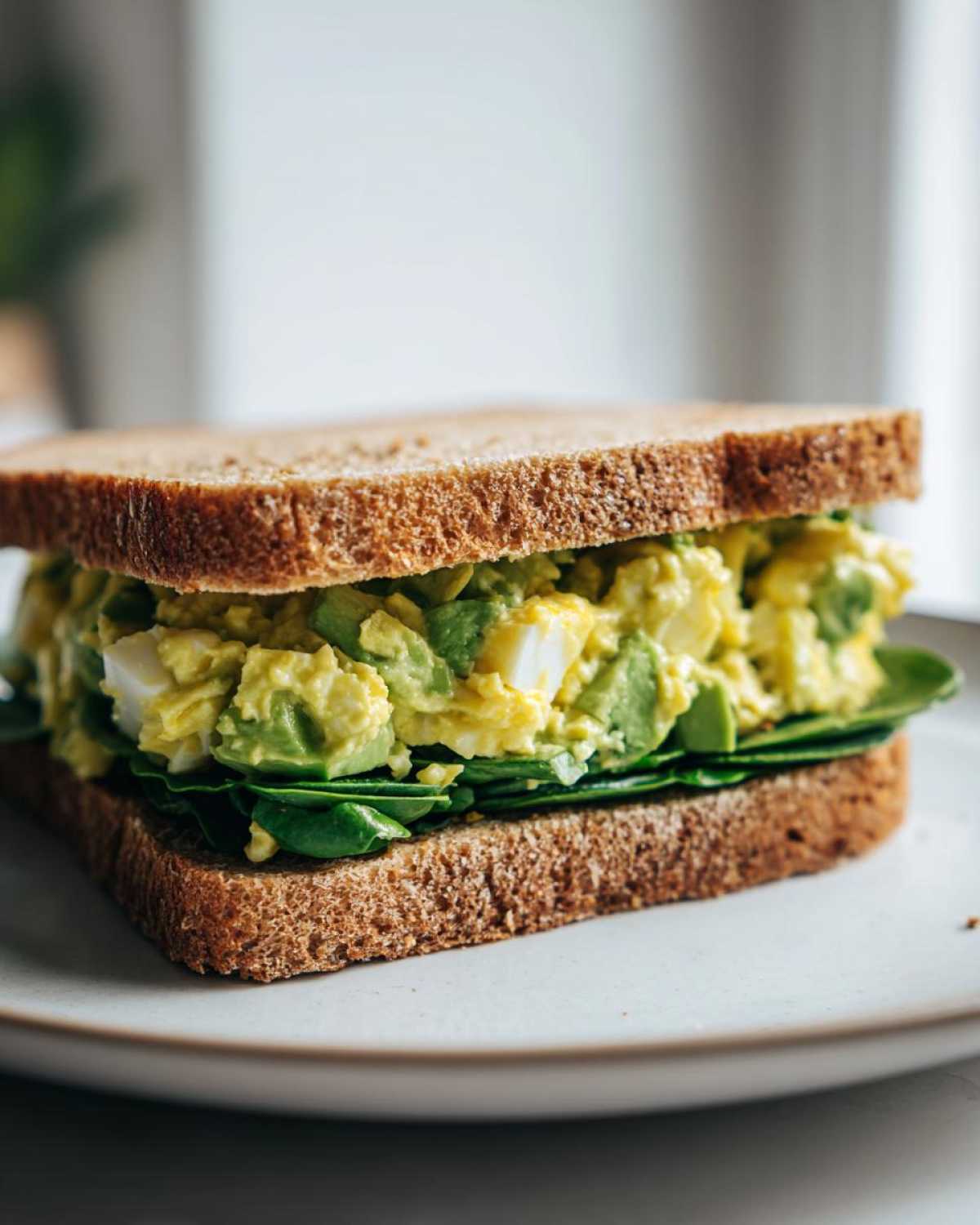 A close-up of an Avocado Egg Salad Sandwich layered with spinach on whole wheat bread, sitting on a light plate.