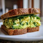 A thick Avocado Egg Salad Sandwich made with whole wheat bread and fresh spinach leaves.