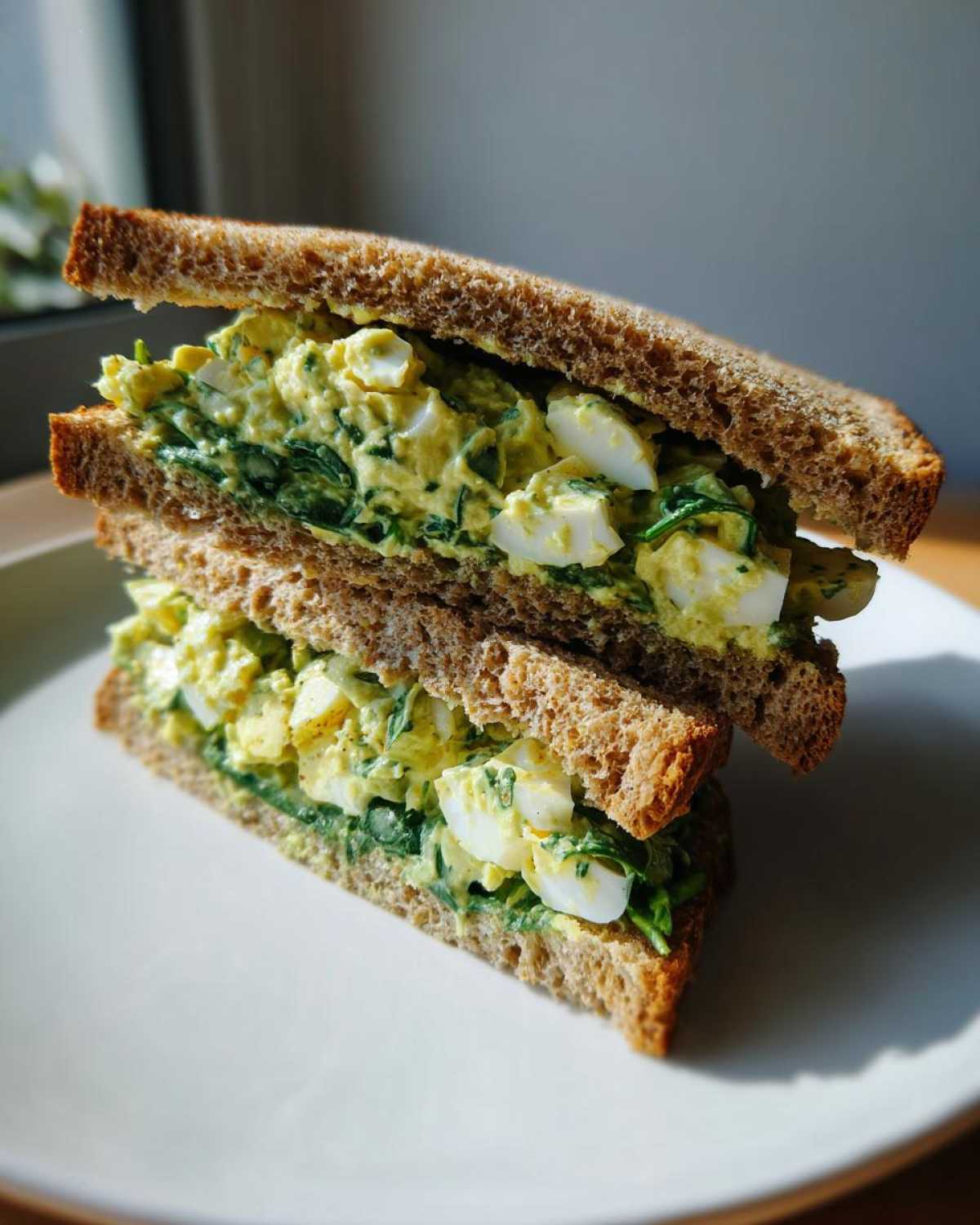 A halved Avocado Egg Salad Sandwich stacked on a white plate, showing creamy filling with egg chunks and spinach on whole wheat bread.