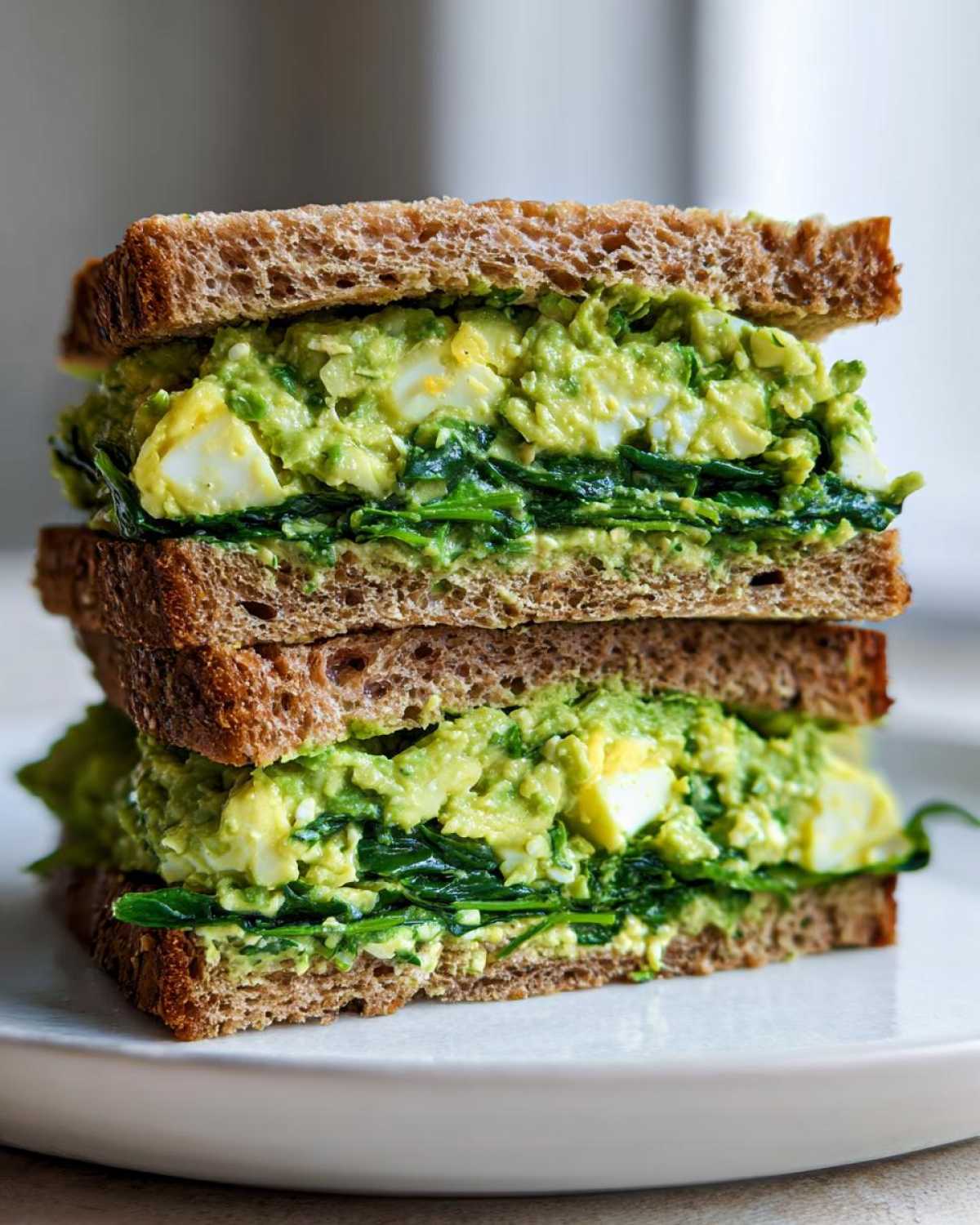 A tall stack of two halves of an Avocado Egg Salad Sandwich made with whole wheat bread and fresh spinach.