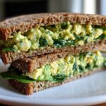 A stacked half of an Avocado Egg Salad Sandwich made with whole wheat bread and fresh spinach.