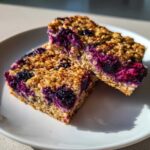 Two golden-brown Blueberry Baked Oatmeal Squares stacked slightly on a white plate, showing juicy berry filling.