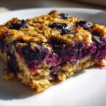 A close-up, sunlit view of one square of moist Blueberry Baked Oatmeal Squares on a white plate.