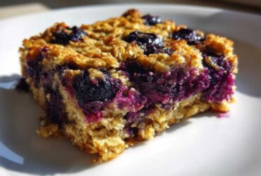 A close-up, sunlit view of one square of moist Blueberry Baked Oatmeal Squares on a white plate.