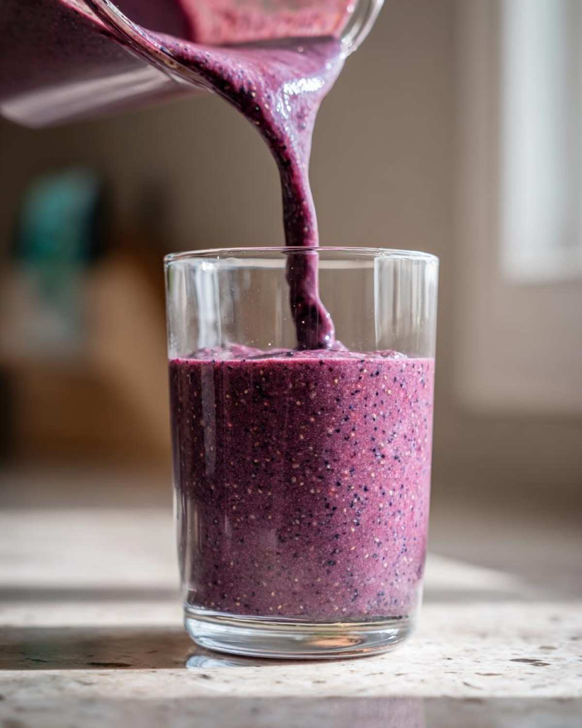 Thick, purple Blueberry Lemon Chia Smoothie being poured from a blender pitcher into a clear glass.