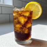 Close-up of a tall glass filled with bubbly, dark Cold Brew Lemonade, ice, and a lemon slice garnish.