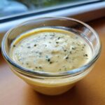 Close-up of creamy, yellow Copycat Olive Garden Salad Dressing speckled with green herbs in a small glass bowl.