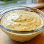 Close-up of creamy, yellow Copycat Olive Garden Salad Dressing with visible herbs in a clear glass bowl.
