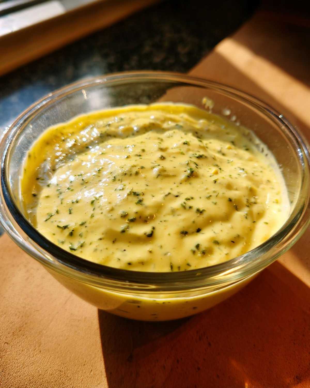 Close-up of creamy, yellow Copycat Olive Garden Salad Dressing speckled with green herbs in a clear glass bowl.